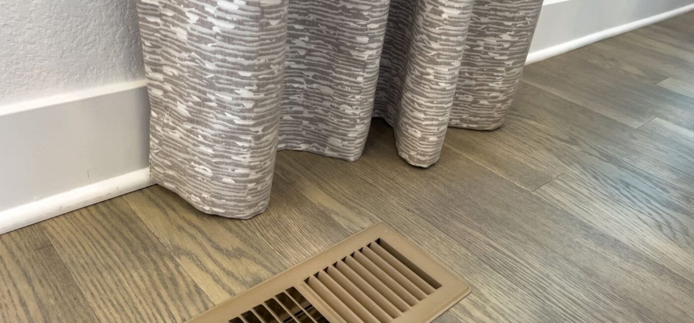 Floor vent for a home heating system next to a curtain and hardwood floor, representing residential furnace airflow and indoor heating.