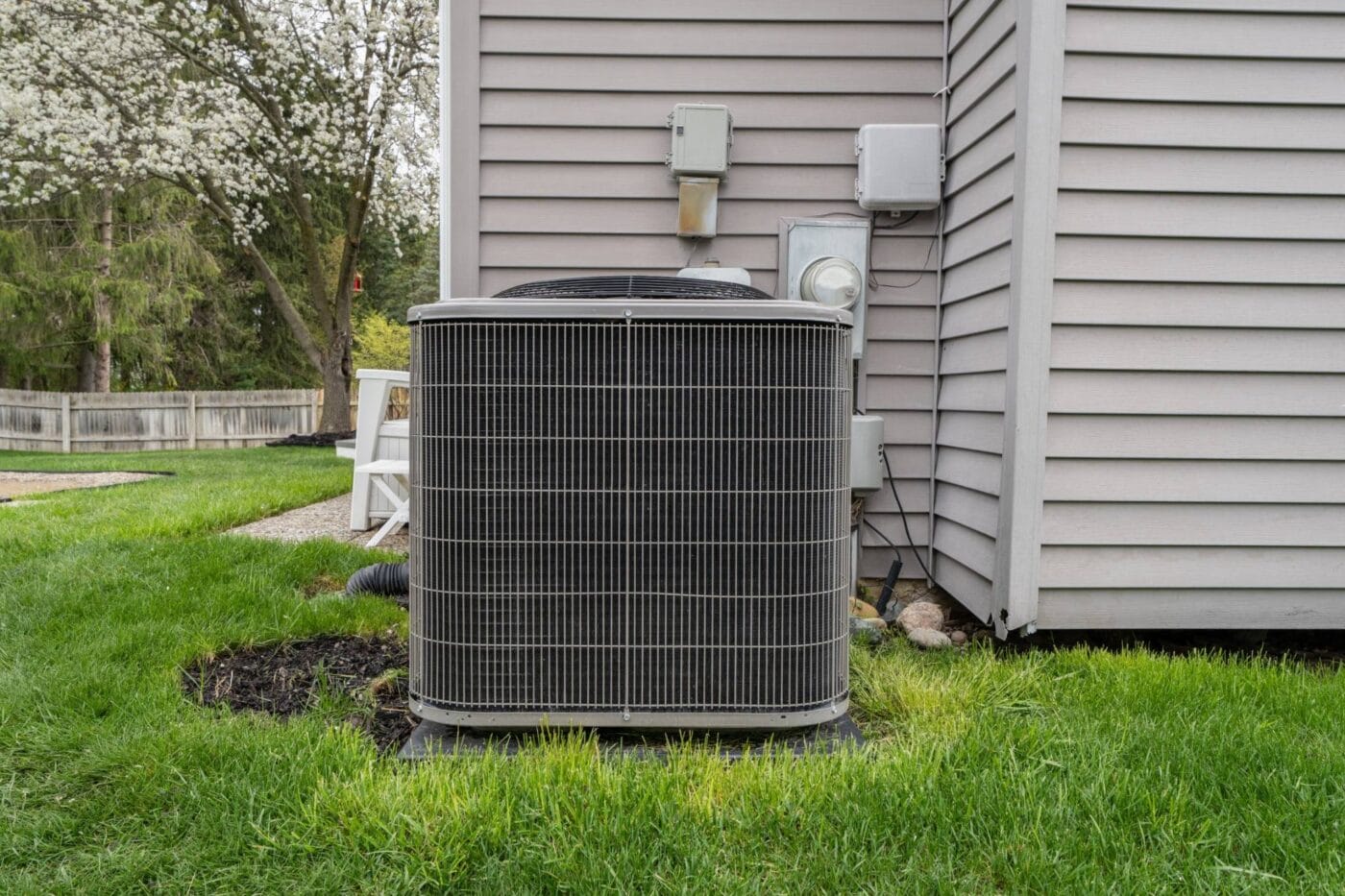 A central air conditioning unit sits on a concrete pad beside a house with vinyl siding, surrounded by green grass and a backyard tree.